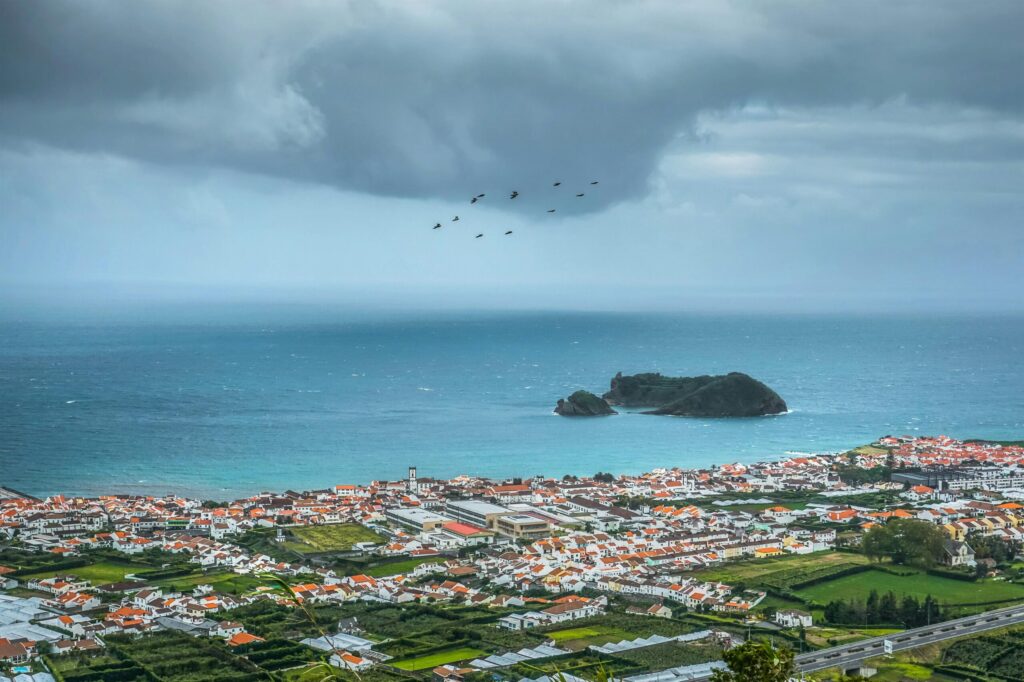 Coastal town with red rooftops beside the ocean and a small offshore island under dramatic storm clouds