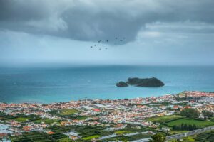 Coastal town with red rooftops beside the ocean and a small offshore island under dramatic storm clouds