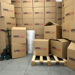 Stacked VANonsite moving boxes in a storage area, with a pallet, hand truck, and packing wrap ready for a move