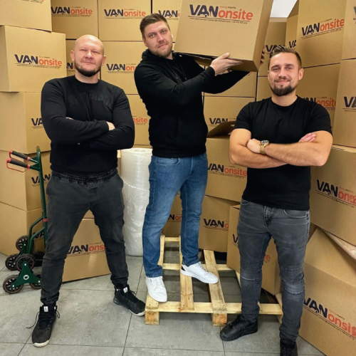 Three VANonsite team members in a warehouse surrounded by branded moving boxes, one holding a box for packing