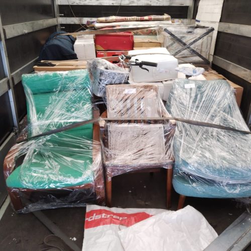 Inside a moving van loaded for an international relocation: three armchairs wrapped in clear plastic film at the front, with stacked cardboard boxes, a wrapped bicycle basket, rolled-up rugs, and other household items secured behind them.