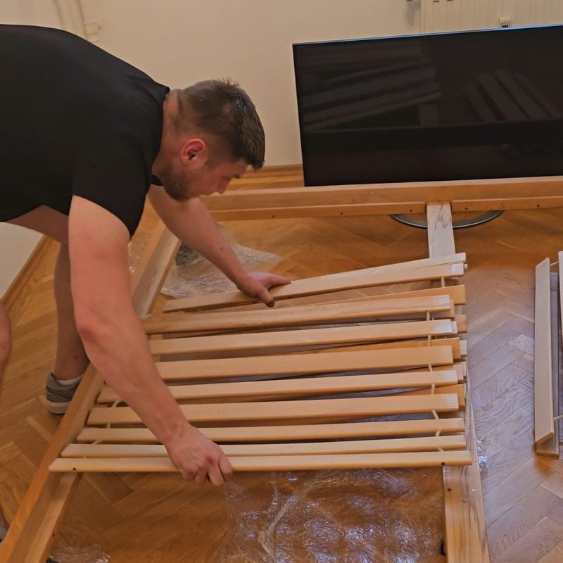 A mover disassembles a wooden bed frame on a parquet floor, preparing furniture for a relocation from Berlin to Budapest.