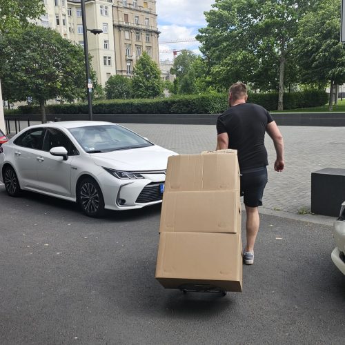 A mover pulls a hand truck loaded with large cardboard boxes along a street during a Berlin to Budapest relocation.