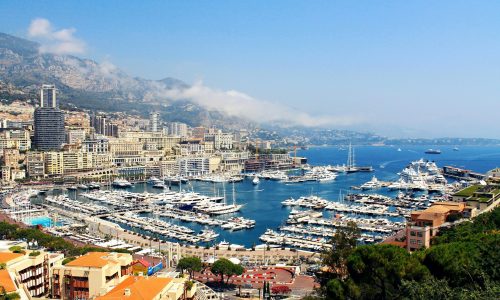Panoramic view of Monaco’s coastline with a crowded marina full of yachts, waterfront buildings, and mountains in the background under a clear blue sky.